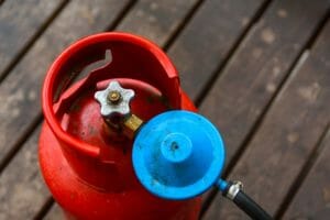 rote Gasflasche ohne Gasflaschenhalterung mit blauem Druckregler auf Holzbohlen