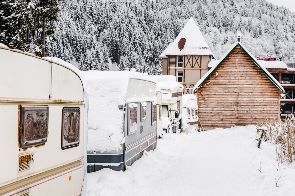 Winterfest gemachte Wohnwagen stehen in einem Skiresort mit Häusern und Wald im Hintergrund 