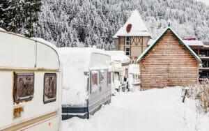 Winterfest gemachte Wohnwagen stehen in einem Skiresort mit Häusern und Wald im Hintergrund